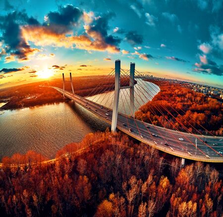 Beautiful panoramic aerial drone view to cable-stayed Siekierkowski Bridge over the Vistula river and Warsaw City skyscrapers, Poland in gold red autumn colors in November evening at sunsetの写真素材
