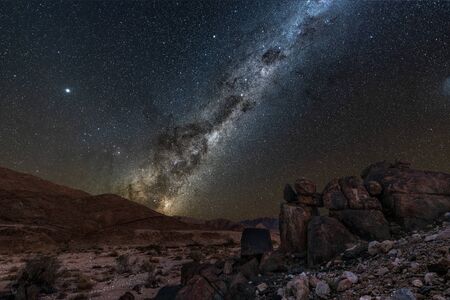 A beautiful night sky landscape with the Milky Way and galactic centre, and interesting rock formations in the foreground and mountains on the horizon, in the Richtersveld National Park, South Africa.の写真素材