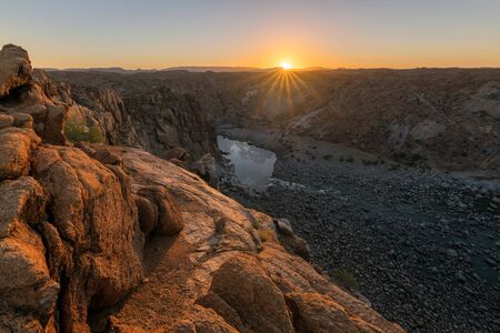 A beautiful landscape view of the Augrabies Falls Gorge, mountains and river in South Africa, taken at sunset with a sunburst on the horizon and rocks in the foreground.の写真素材