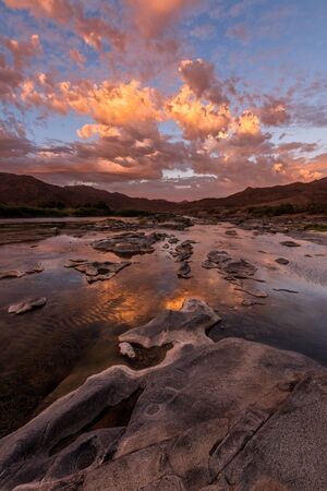 A vertical landscape of a golden sunset over the mountains and calm waters of the Orange River, with dramatic orange clouds reflecting in the waterâs surface, taken in the Richtersveld South Africa.の写真素材