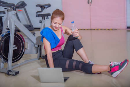 An athletic woman in a sports suit was sitting on the floor with a bottle of water and used a laptop at the gym. Sports and recreation concepts.の写真素材