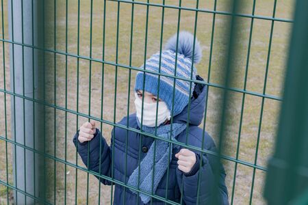 A small boy in a medical mask stands behind a metal fenceの写真素材