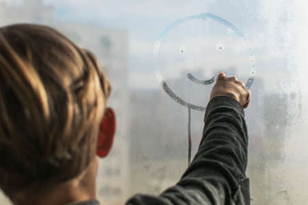 A little boy draws a smiley face on a fogged, wet windowの写真素材