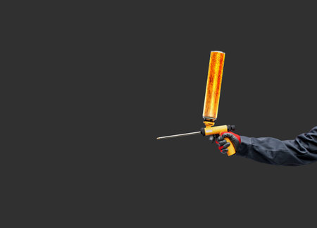 A worker's hand holds a polyurethane foam expanding glue gun on a dark background