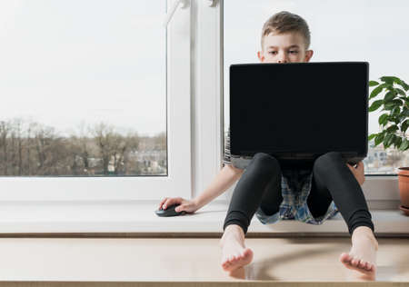 A small boy sits at a computer near the window in the apartmentの写真素材