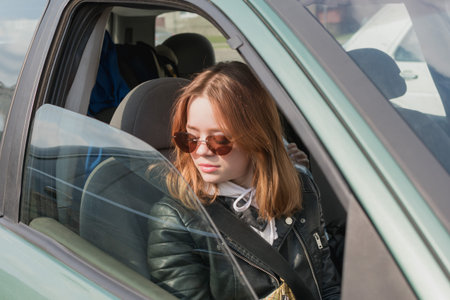 A young girl wearing glasses and a leather jacket is sitting in a car