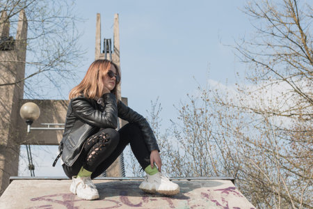A young girl wearing glasses and a leather jacket walks in the park near the suspension bridge
