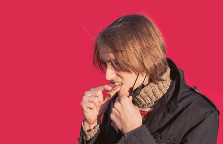 A young guy with long hair in a protective medical mask eats sausage