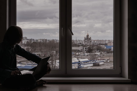 A young guy is sitting near the window with a laptopの写真素材