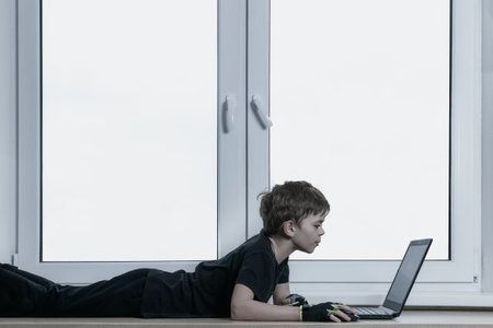 A little boy is sitting by the window and playing on the computer