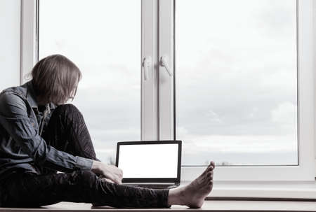 A young guy is sitting near the window with a laptopの写真素材