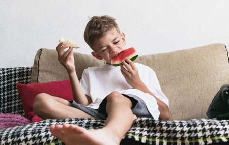 Little boy eats watermelon and melon sitting on the couchの写真素材