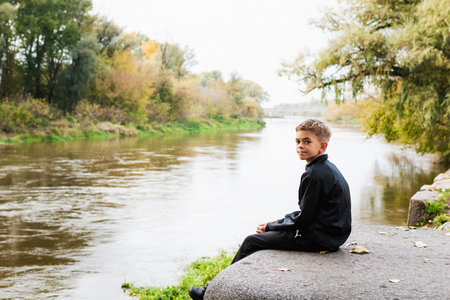 A little boy sits thoughtfully on the bank of the riverの写真素材