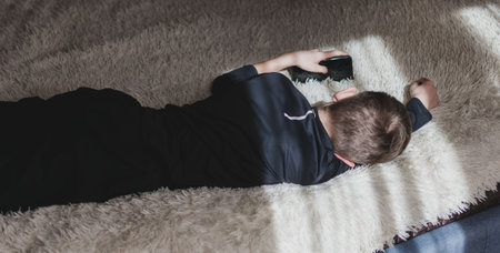A little boy is lying on the couch and playing with his phone, The sun is shining through the window