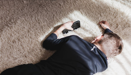 A little boy is lying on the couch and playing with his phone, The sun is shining through the window