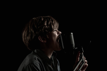 Portrait of a little boy with long hair singing near a microphone on a dark backgroundの写真素材
