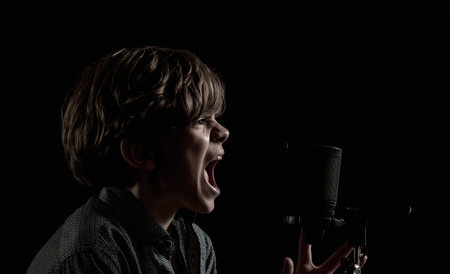 Portrait of a little boy with long hair singing near a microphone on a dark backgroundの写真素材