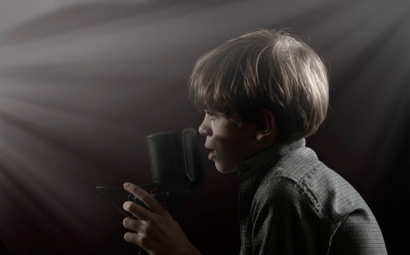 Portrait of a little boy with long hair singing near a microphone on a dark backgroundの写真素材