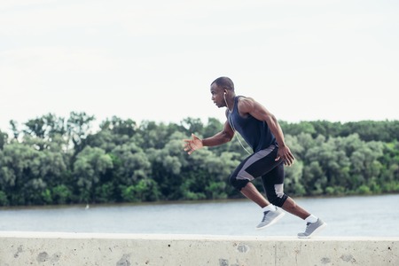 Attractive fit man running fast at sunset light, black man doing workout outdoors, athletic runner in windbreaker jogging roadの写真素材