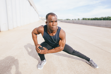 Cropped shot of male runner in black sportswear stretching legs before doing morning workout. Young African jogger warming up before outdoor run, standing on steps of concrete stair, touching toesの写真素材