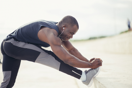 Cropped shot of male runner in black sportswear stretching legs before doing morning workout. Young African jogger warming up before outdoor run, standing on steps of concrete stair, touching toesの写真素材