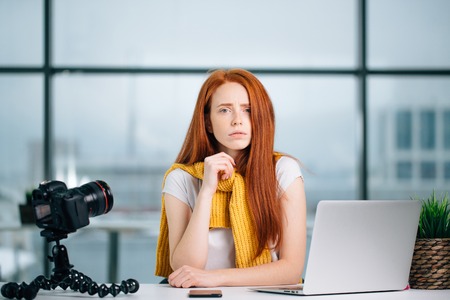 sad redhead girl vlogger sitting at table with laptop and looking at camera.の写真素材