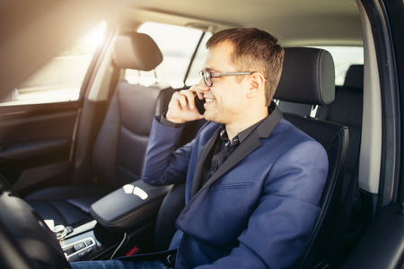 Handsome businessman talking with phone sitting with laptop on backseat of carの写真素材