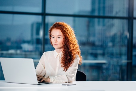 redhead businesswoman sitting at bright modern work station and typing on laptopの写真素材