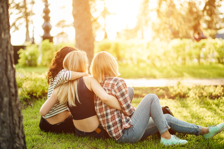 Back view of three beautiful women sitting on grass and hugsの写真素材