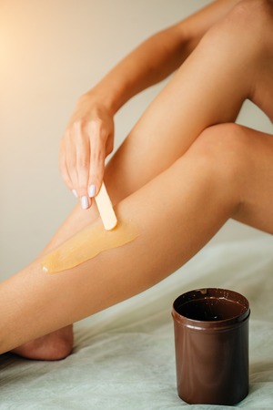 Young woman depilating legs with wax sitting on bedの写真素材