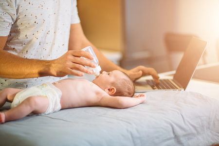 dad trying to work while standing with his newborn babe in home office interiorの写真素材