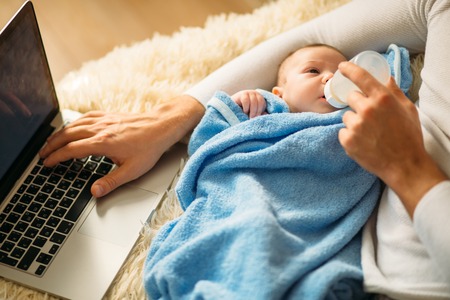 man feeding little baby while lying on bed and working on laptopの写真素材