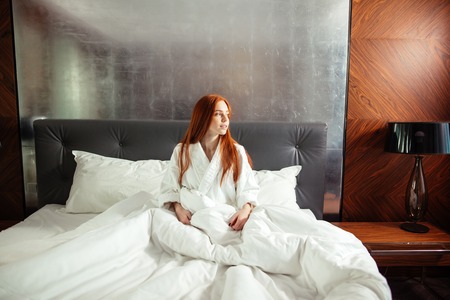 redhead young woman with freckles sitting on bed relaxingの写真素材