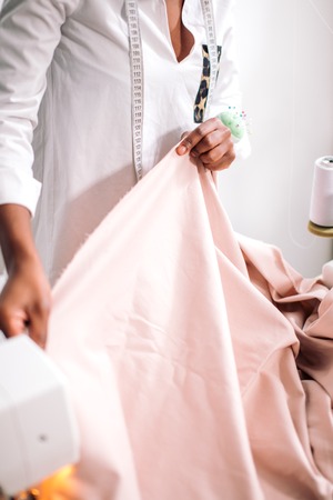 Young african woman seamstress looking at pink fabrics and standing in workshopの写真素材