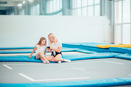 Instuctor and a girl relaxing after workout sitting on mat, using phone in gymの写真素材