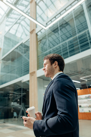 Confident man holding coffee cup and using his smart phone while walking in hallの写真素材