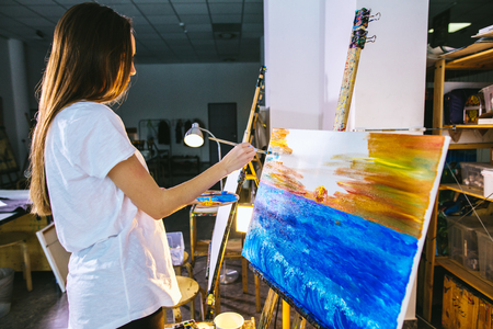 Woman artist painting a picture on easel with oil paints in her workshopの写真素材