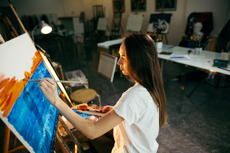 Woman artist painting a picture on easel with oil paints in her workshopの写真素材