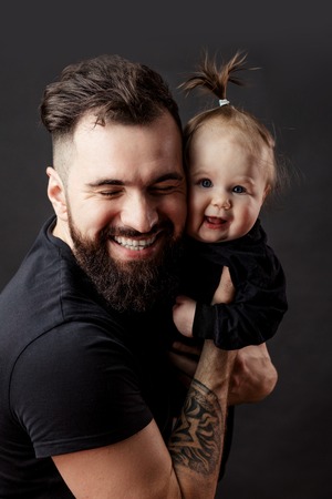 Handsome tattooed young man holding cute little baby on black backgroundの写真素材