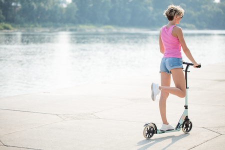 cute female rider in white shoes is riding on blue kick scooter on road in park at sunny dayの写真素材