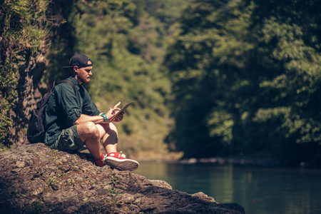 Single male hiker using tablet on nature while sitting on rocky river shoreの写真素材