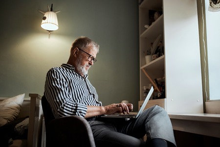 mature bearded man working from home with laptop. sitting at desk near windowの写真素材