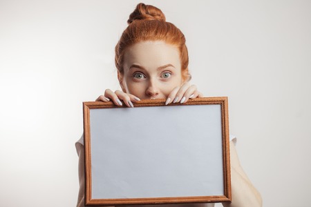 Portrait of cheerful redhead girl with hair bun holding empty wooden frame.の写真素材