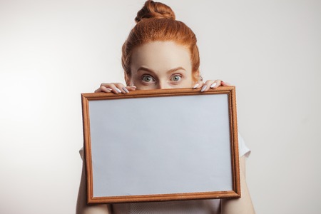 Portrait of cheerful redhead girl with hair bun and freckles holding empty wooden frame.の写真素材