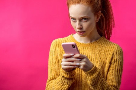 Portrait of cute ginger girl using smartphone isolated on pink bの写真素材