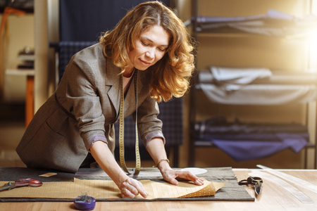 Professional adult female tailor marking cloth pattern with chalk at sewing workshop and looking at cameraの写真素材