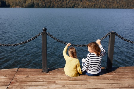 rear view mother and daughter sitting on pier warm autumn dayの写真素材