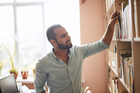 Young Arabian bearded male student choosing book between shelves in the libraryの写真素材