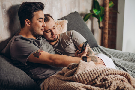 Happy gay couple lying together in bed and using smartphone, looking at photosの写真素材