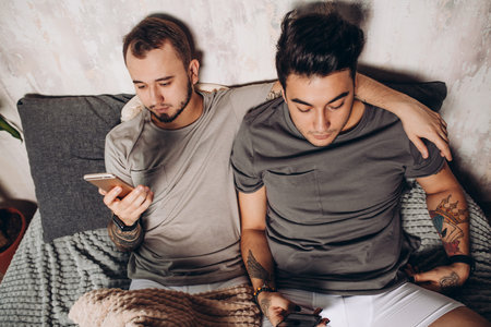 Happy gay couple lying together in bed and using smartphone, looking at photosの写真素材
